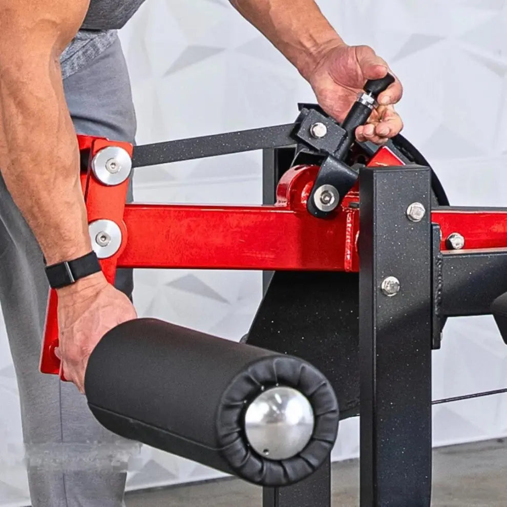 Man adjusting the pin on a red and black selectorized prone leg curl machine with padded leg roller and adjustment straps.