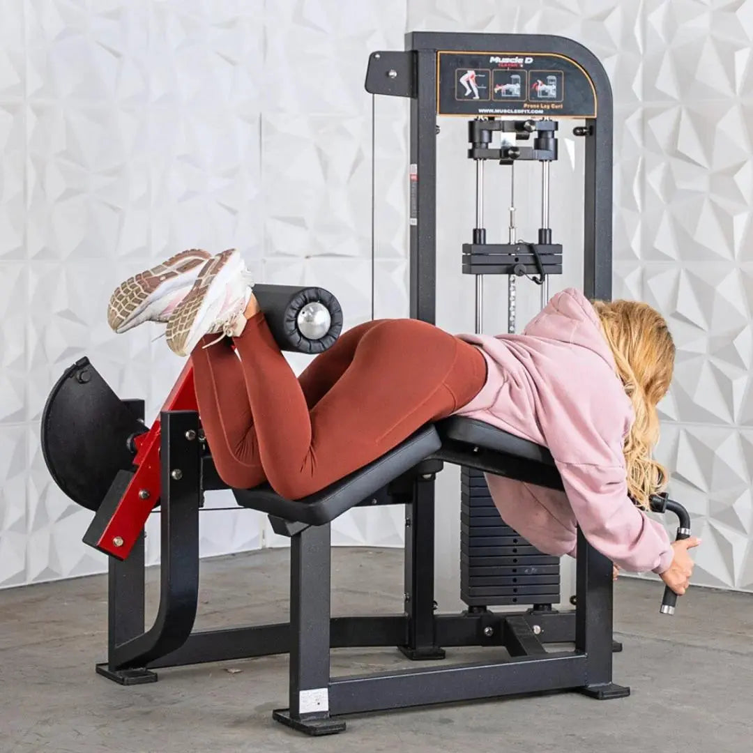 Woman performing a prone leg curl on a black Muscle D Fitness selectorized leg curl machine with red padding and weight stack visible in the background.