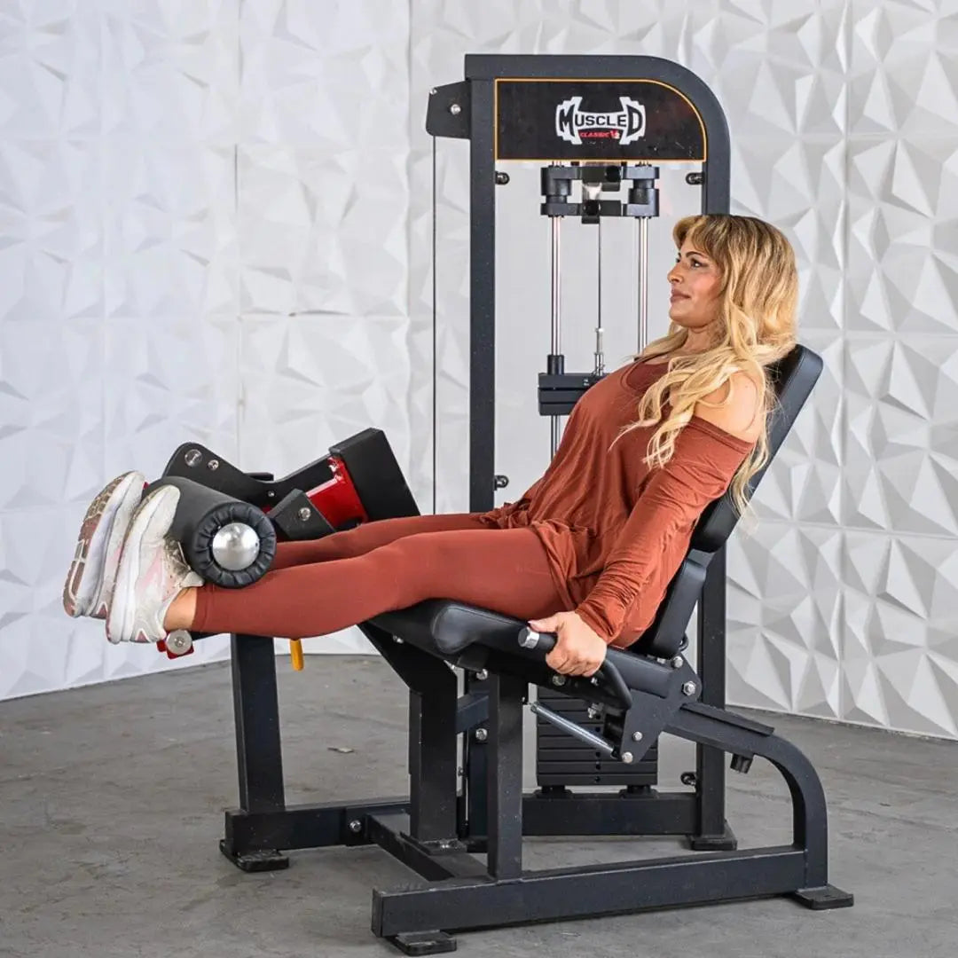 Woman performing seated leg curl on a black and white selectorized leg curl machine with orange accents in a gym setting.