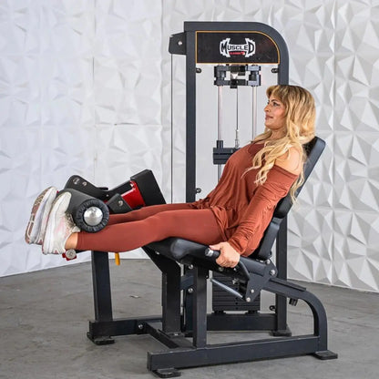 Woman performing seated leg curl on a black and white selectorized leg curl machine with orange accents in a gym setting.
