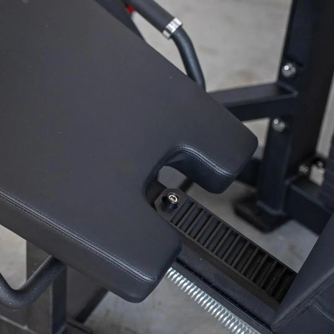 Close-up of black padded leg curl machine seat and footplate with ribbed texture, showing adjustment mechanism and chrome-handled lever on dark frame.