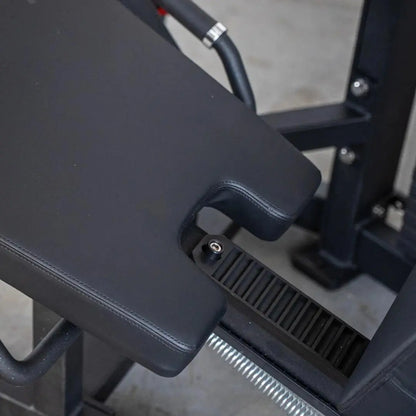 Close-up of black padded leg curl machine seat and footplate with ribbed texture, showing adjustment mechanism and chrome-handled lever on dark frame.