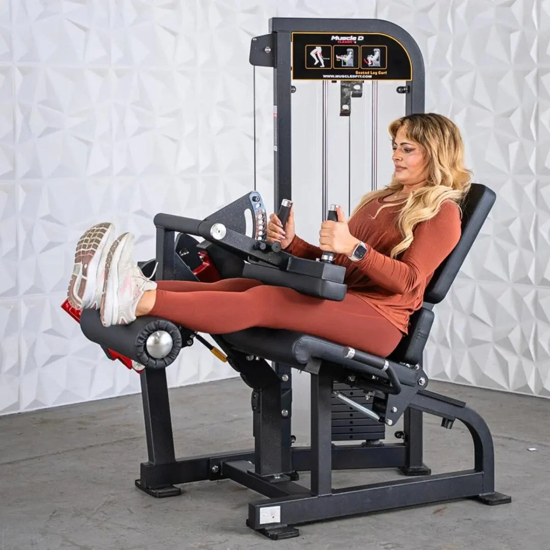 Woman performing seated leg curl on a black selectorized leg curl machine with weight stack and instruction placard on the backrest.