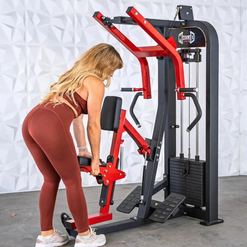 Woman performing a seated row exercise on a commercial machine with red arms, black frame, and footrests, against a white textured backdrop.