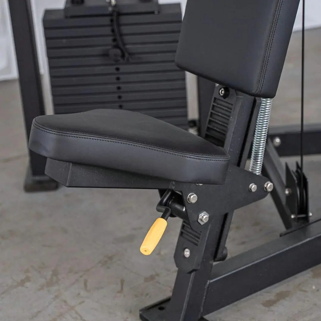 Close-up of a shoulder press machine seat with black padded upholstery, vertical back support, and adjustable height lever with yellow handle. Stacked weight plates visible in the background.