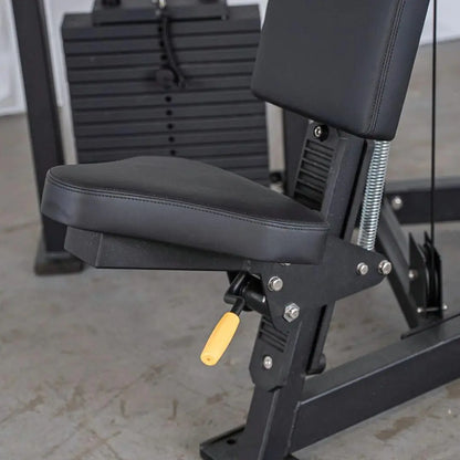 Close-up of a shoulder press machine seat with black padded upholstery, vertical back support, and adjustable height lever with yellow handle. Stacked weight plates visible in the background.