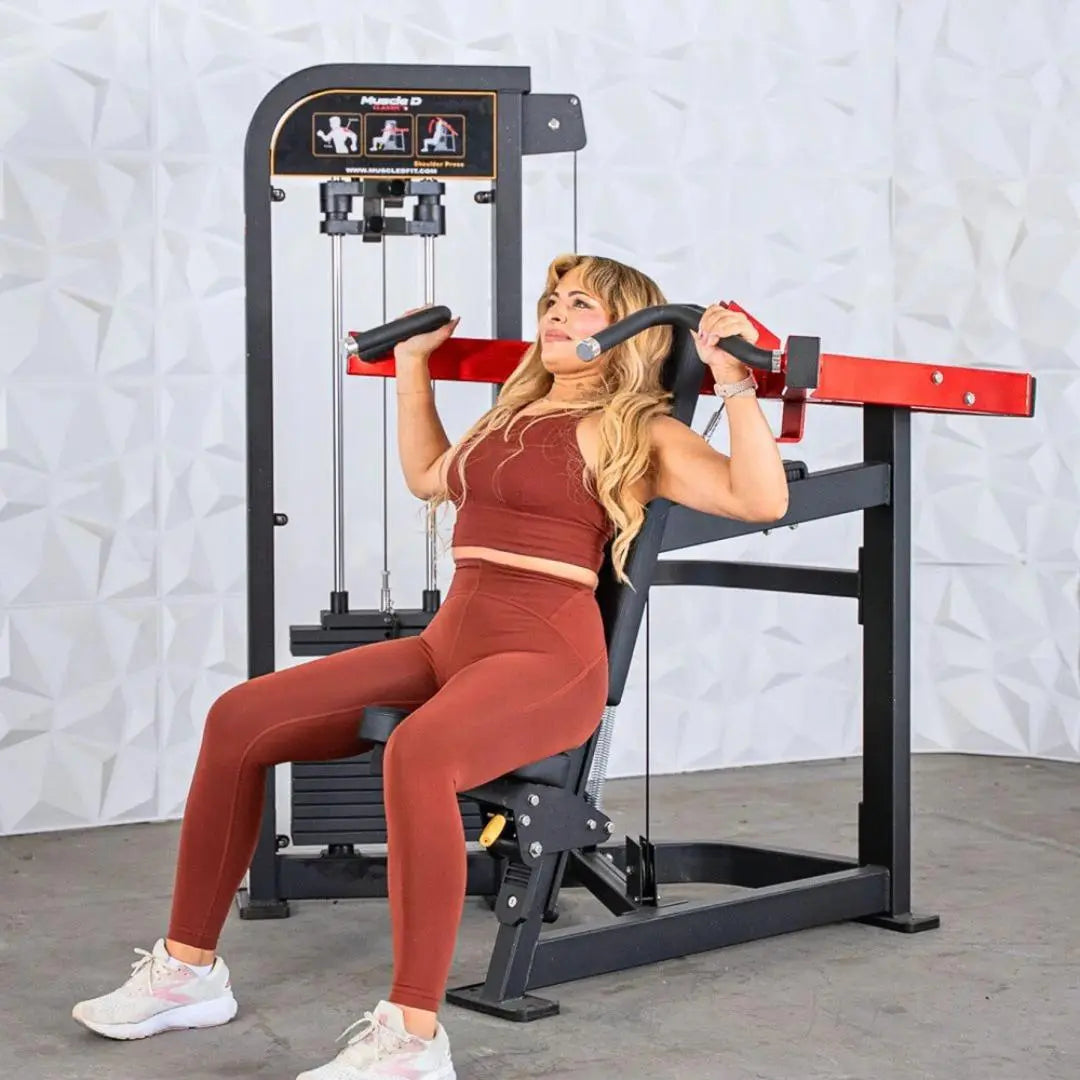 Woman performing shoulder press on a black and red selectorized shoulder press machine with red handles and weight stack cables visible.