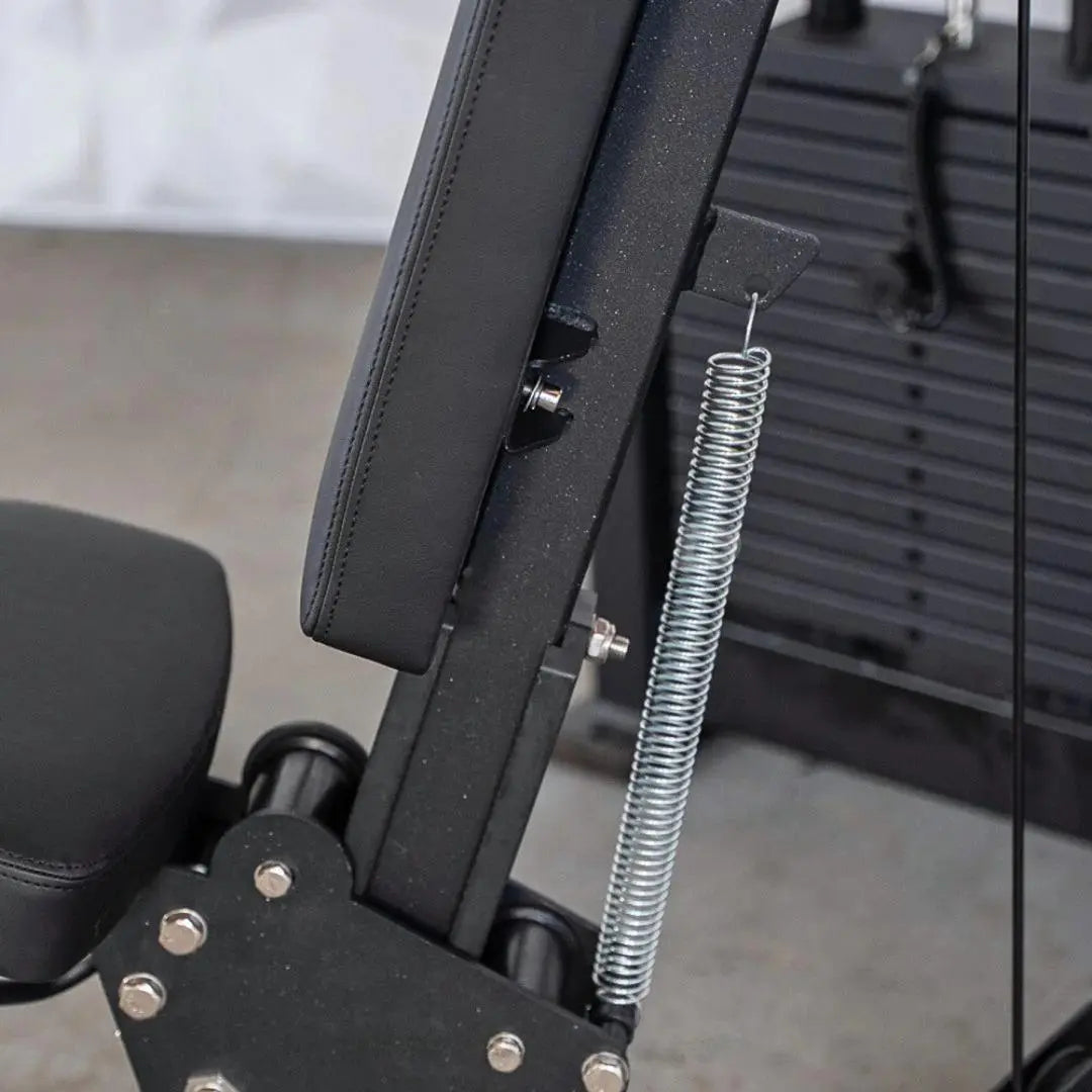 Close-up of shoulder press machine showing black padded seat back, adjustment lever, and silver coiled spring mechanism on the frame.