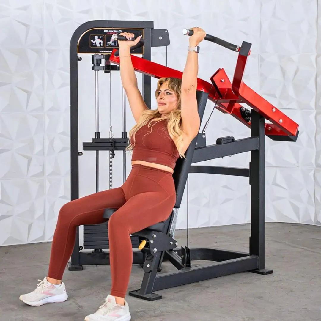 Woman performing shoulder press on a black and red selectorized shoulder press machine with instruction graphics visible on the overhead panel.