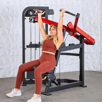 Woman performing shoulder press on a black and red selectorized shoulder press machine with instruction graphics visible on the overhead panel.