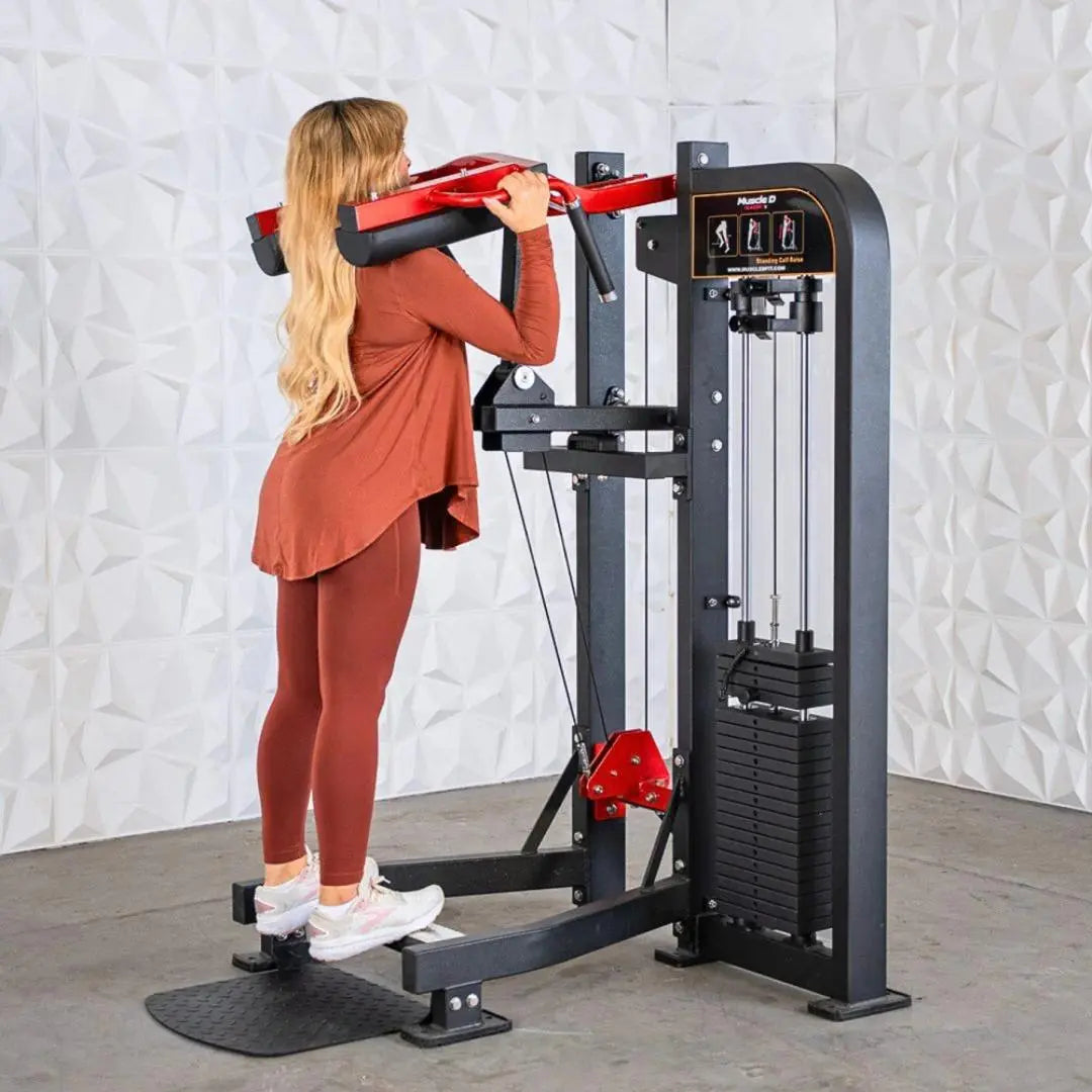 Woman performing a standing calf raise on a black selectorized machine with red handles and weight stack, against a textured white wall.