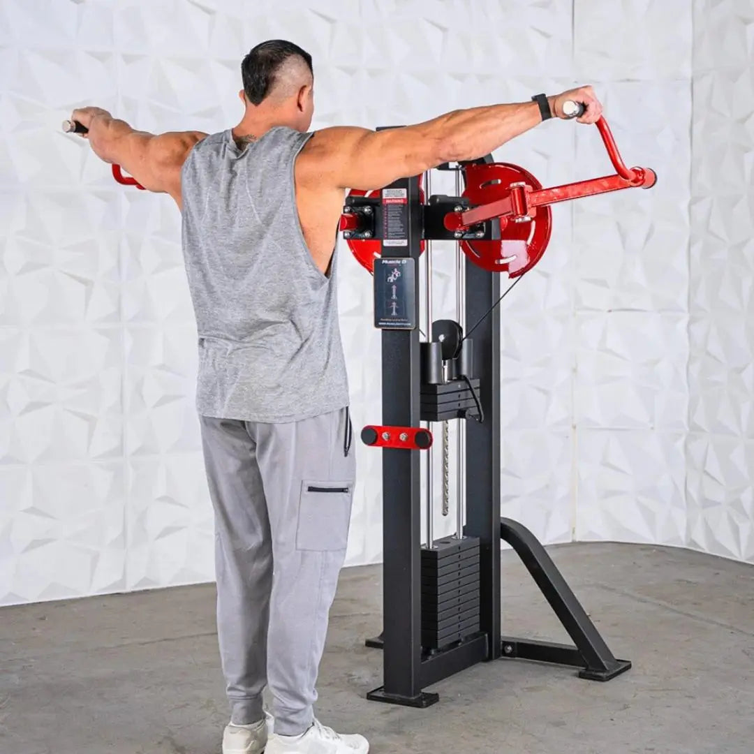 Man performing a standing lateral raise on a selectorized machine with red handles, black frame, and weight stack, against a white brick background.
