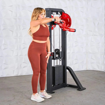 Woman performing a lateral raise on a selectorized standing lateral raise machine with red handles and black frame against a white brick background.