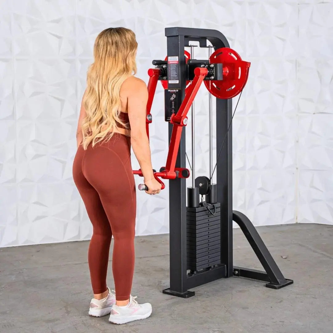 Woman performing a lateral raise on a black and red selectorized machine with weight stack. The machine features red articulating arms and is set against a white brick wall.