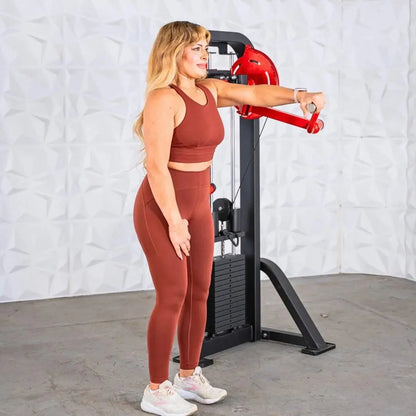 Woman performing a standing lateral raise on a black and red selectorized machine with padded handles and weight stack visible in the background.