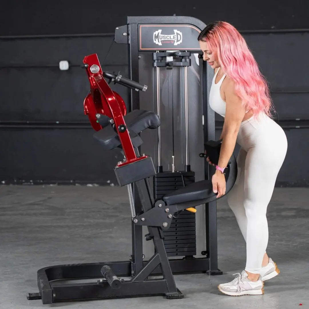 Woman performing a triceps extension exercise on a black and red selectorized triceps machine in a gym with dark walls.