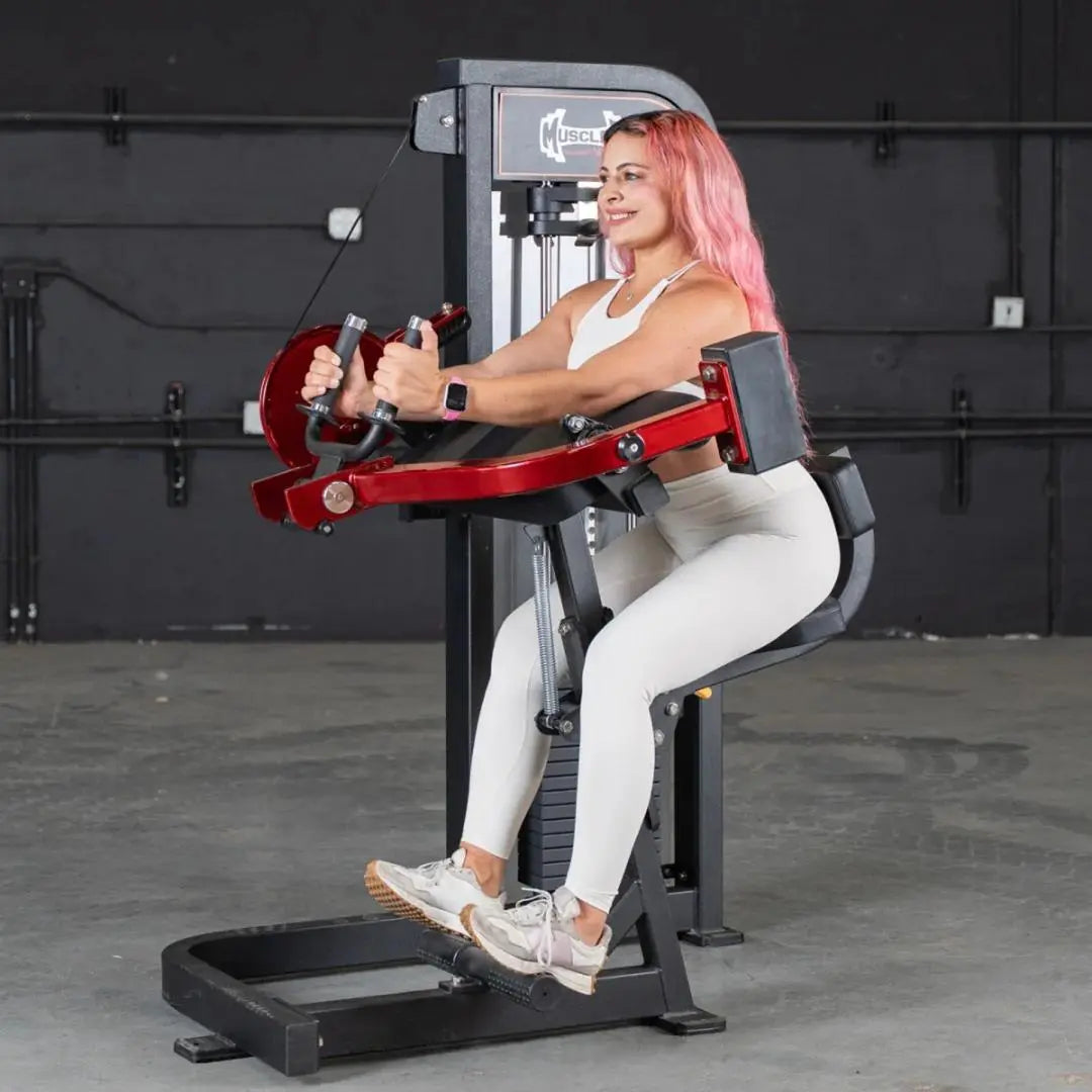 Woman performing a triceps extension on a red and black selectorized machine in a gym with dark walls and concrete flooring.