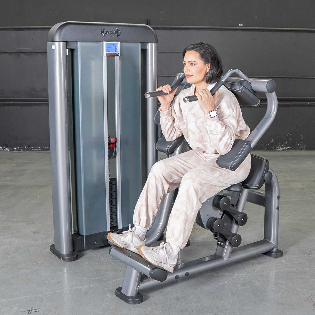 Woman using Muscle D shoulder press machine in a beige tracksuit.