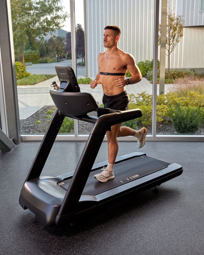 Shirtless man wearing a chest strap runs on a treadmill in a bright gym.