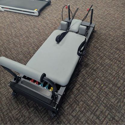 Gray Pilates reformer with padded bench and resistance cords.