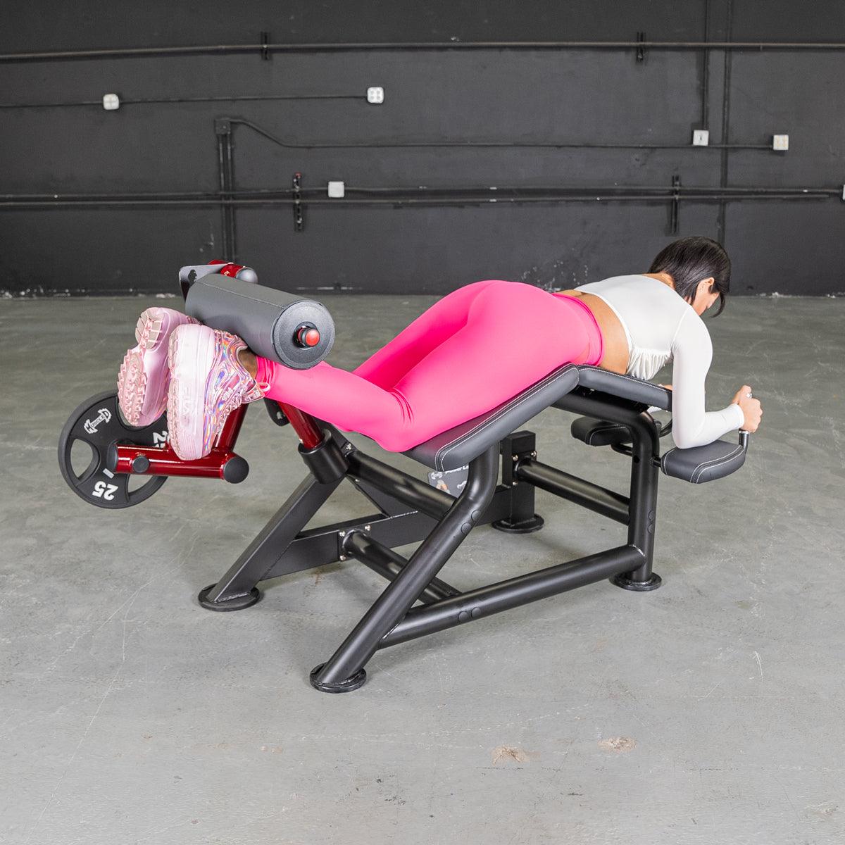Woman wearing pink leggings using a prone leg curl machine at the gym.