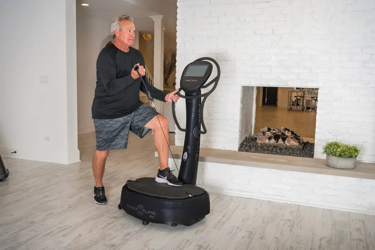 Man performing a dynamic exercise on a black Power Plate vibration platform with digital display, in a modern home gym with white brick walls.