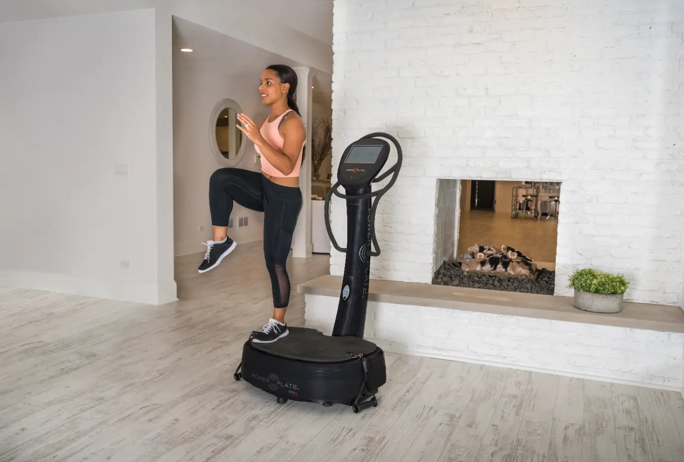 Woman performing a high knee exercise on a black Power Plate vibration platform in a modern home gym with white brick walls.