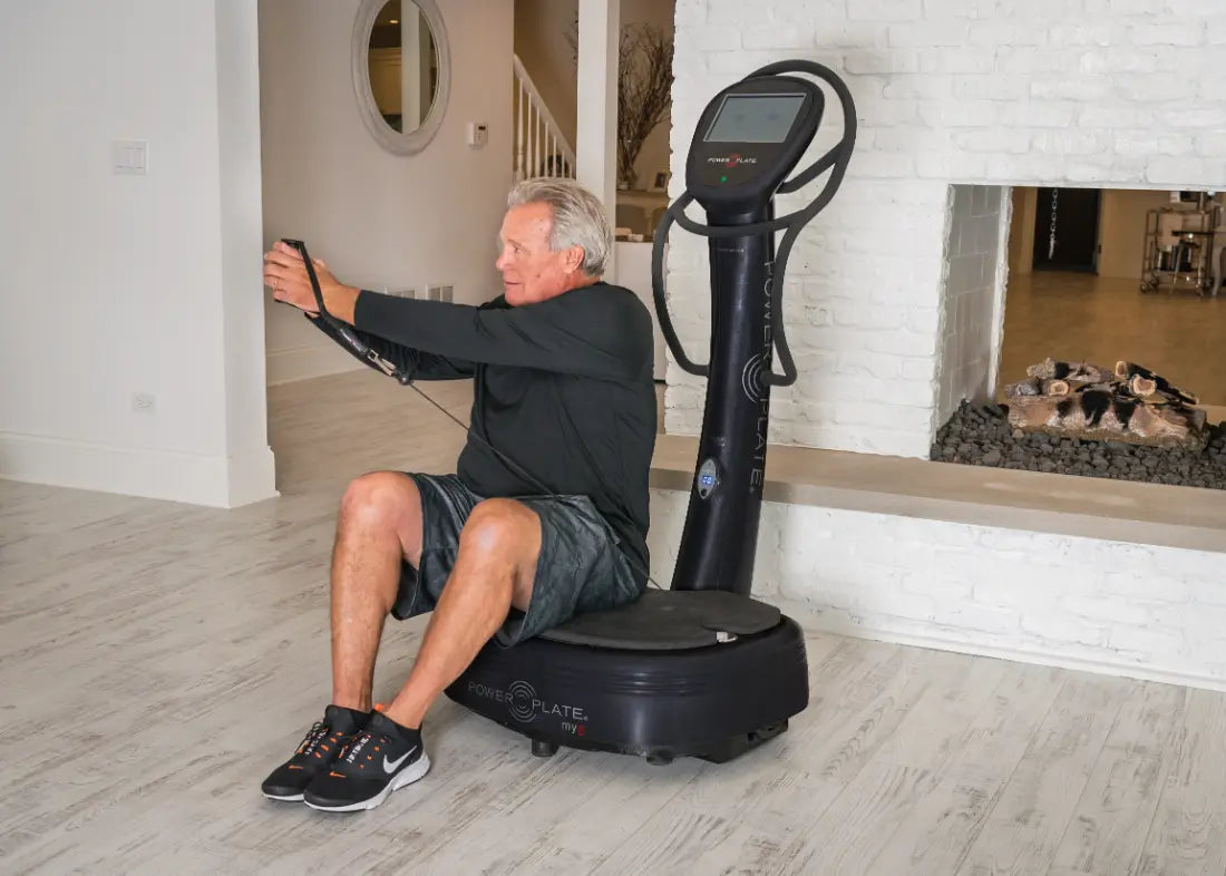 Man performing a seated exercise on a black Power Plate vibration platform with digital display, holding resistance bands in a home gym setting.