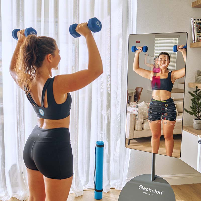 A woman in a black sports bra and shorts is lifting dumbbells while looking at her reflection in the Echelon Reflect 50 Smart Fitness Mirror, set in a bright room with sheer curtains and a yoga mat nearby.