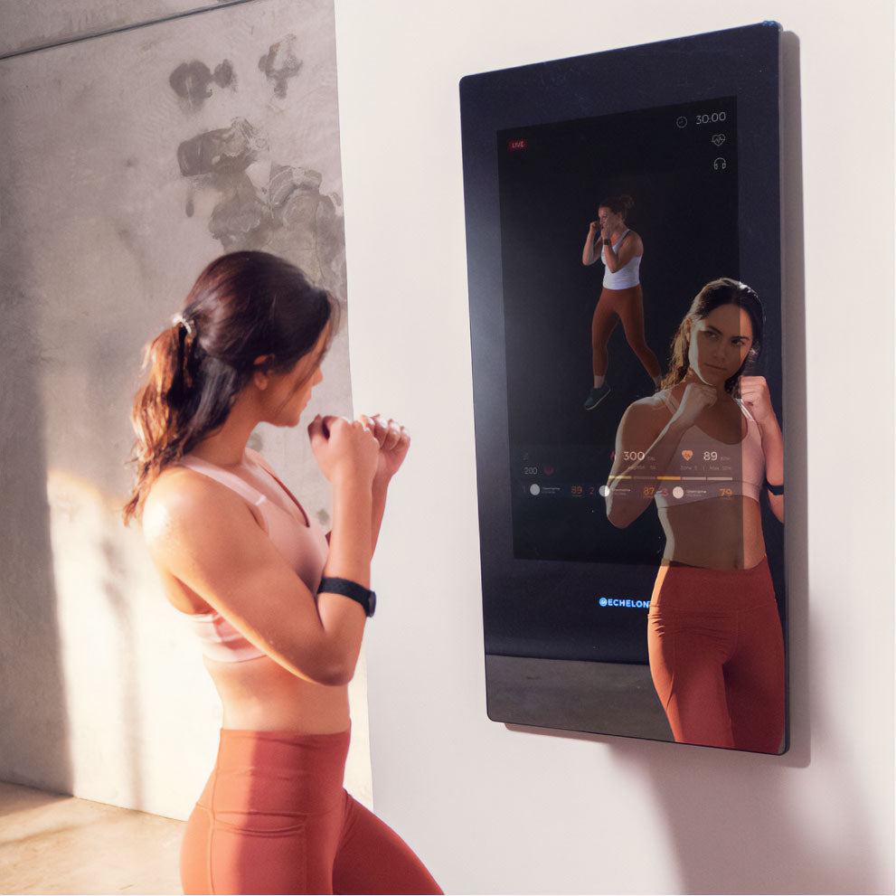 A woman in athletic wear engages in a workout while using the Echelon Reflect 50 Smart Fitness Mirror, which displays workout metrics and a live video feed.