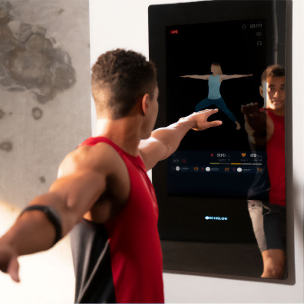 A man in a red tank top interacting with the Echelon Reflect 50\ Smart Fitness Mirror, which displays a fitness instructor demonstrating a yoga pose.