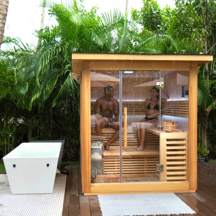 Outdoor wooden sauna with glass doors, two people relaxing inside amid tropical greenery.