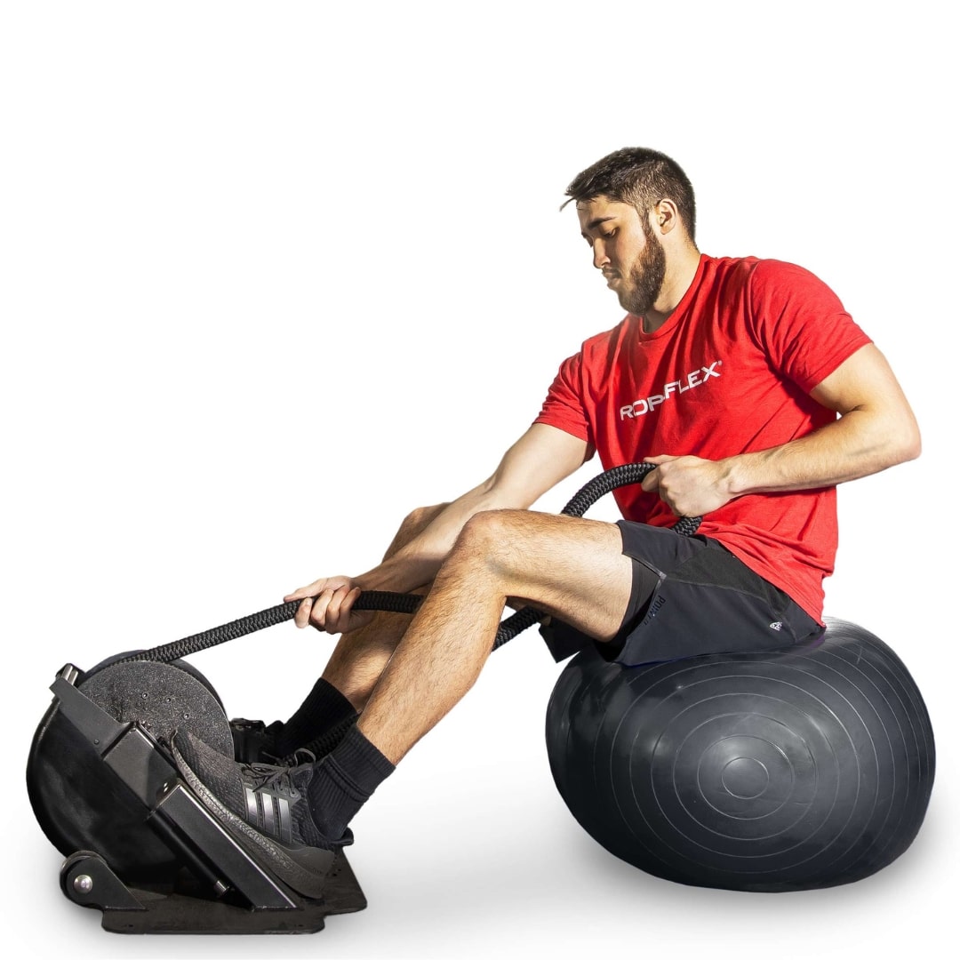 Man exercising with RopFlex machine on black stability ball, wearing red shirt and black shorts.