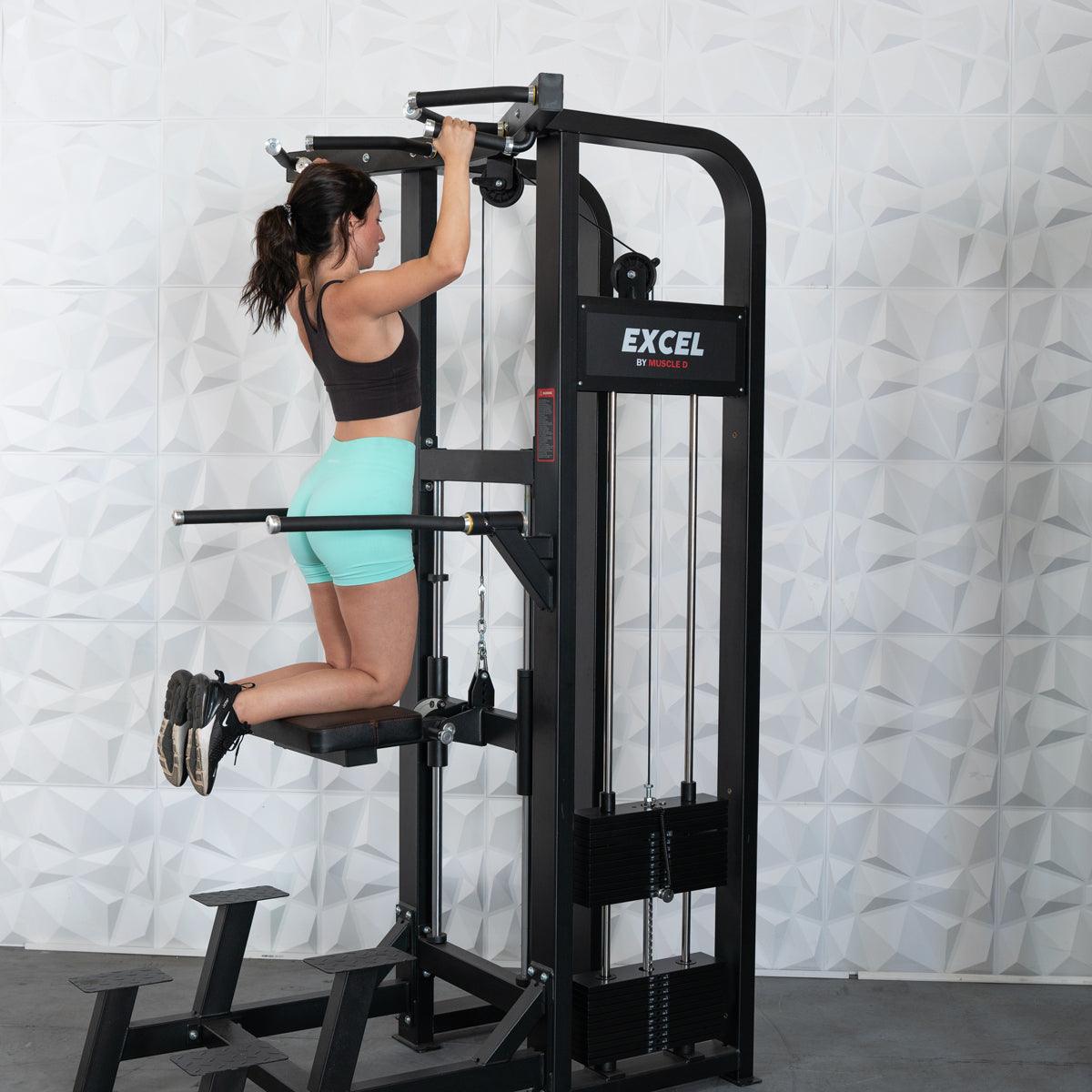Woman using Excel pull-up machine in a gym, wearing teal shorts.