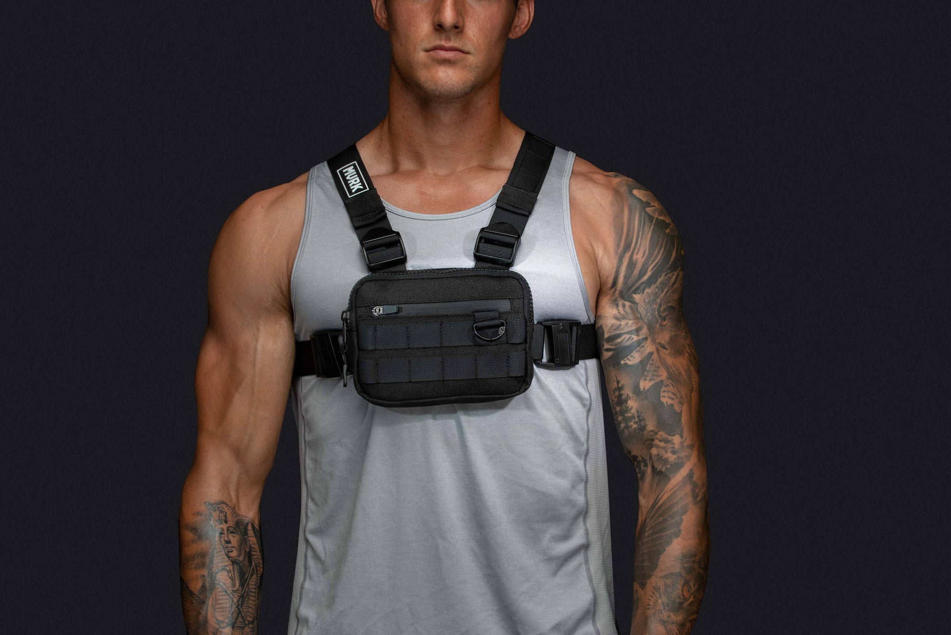 Athletic man in gray tank top wearing a black chest rig with straps.