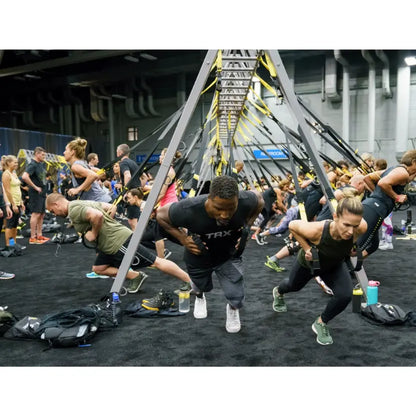 Multiple people performing suspended exercises on a TRX suspension trainer system with yellow and black straps attached to an elevated gray truss frame in a gym setting.