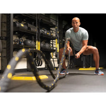 Man performing battle rope exercise with black and yellow rope in a gym with equipment storage racks in the background.