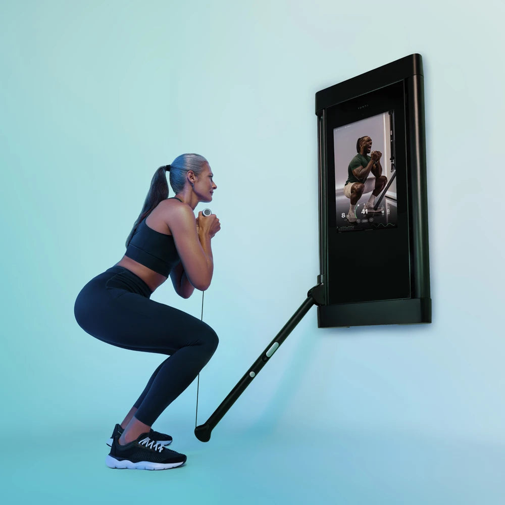 Woman doing a squat with a resistance band attached to a wall-mounted Tonal trainer.