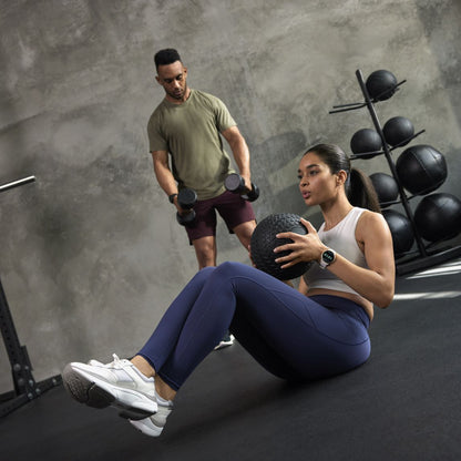 A woman in athletic wear performs a medicine ball exercise while wearing a Garmin Vivoactive 5, as a man in the background lifts dumbbells in a gym setting.