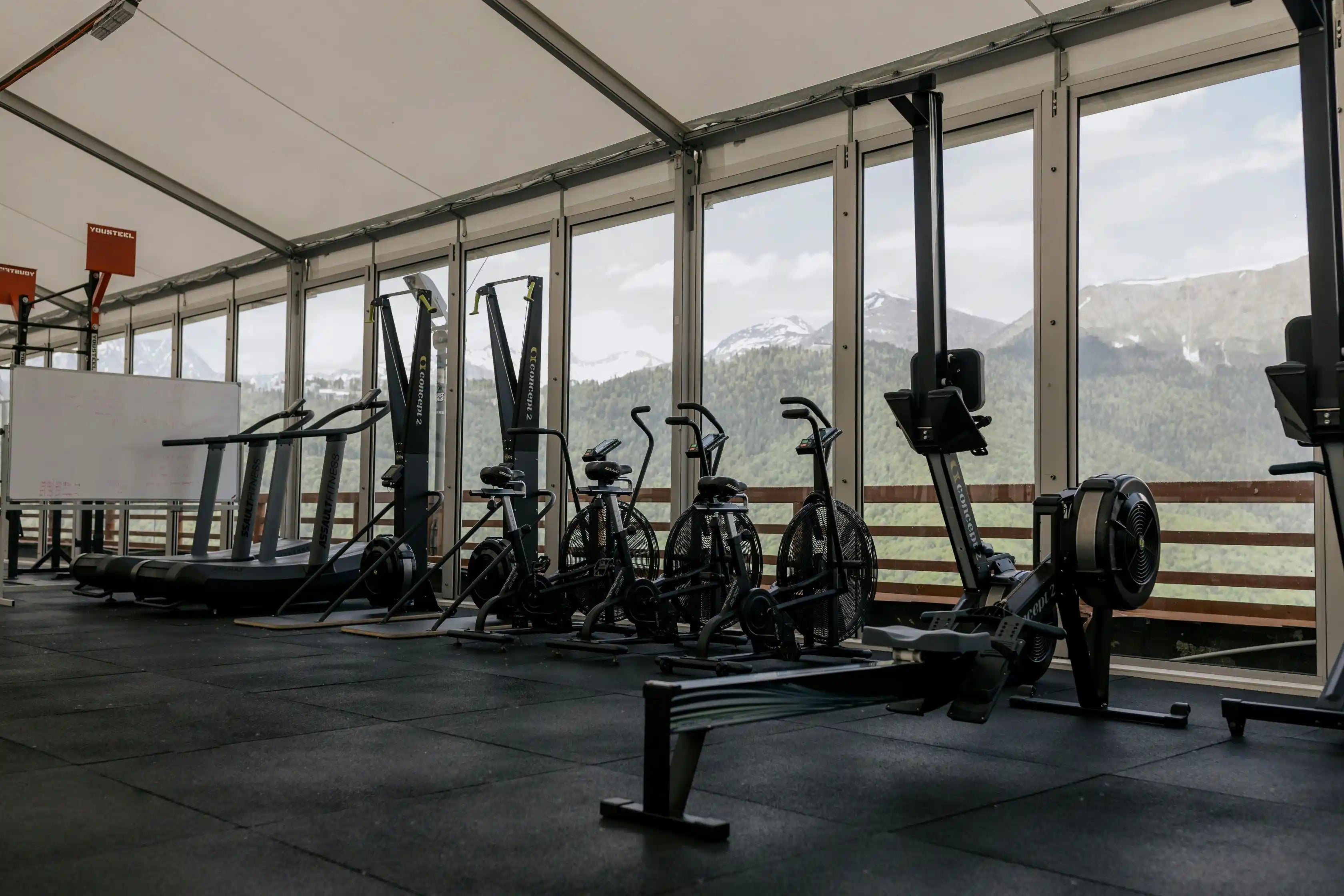 Gym interior with exercise equipment and large windows overlooking a mountainous landscape.