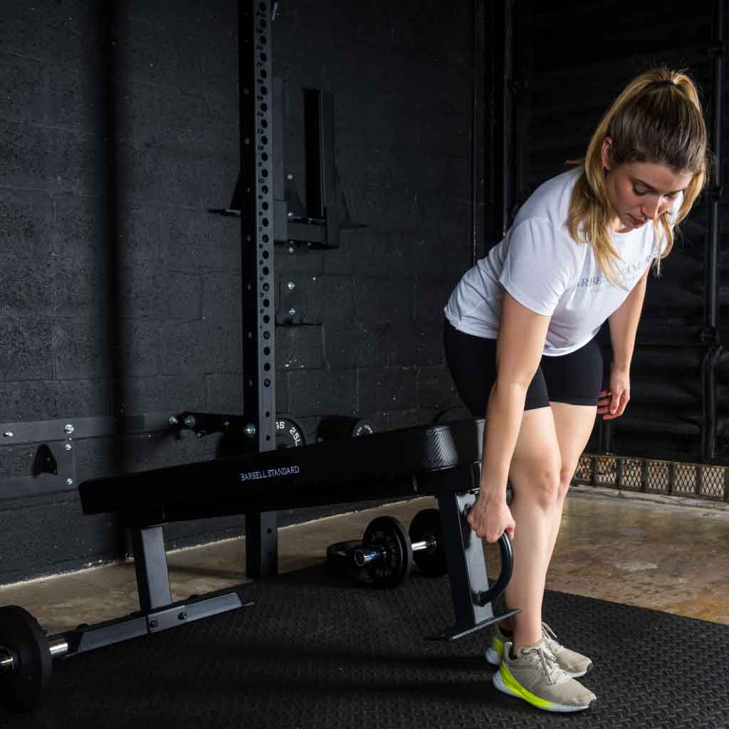 Woman in a gym bending to lift a weight plate near a bench.