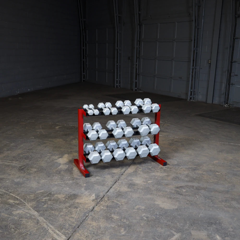 A rack of hexagonal dumbbells in various weights, arranged neatly on a red and black stand, placed in an empty gym space with a concrete floor.