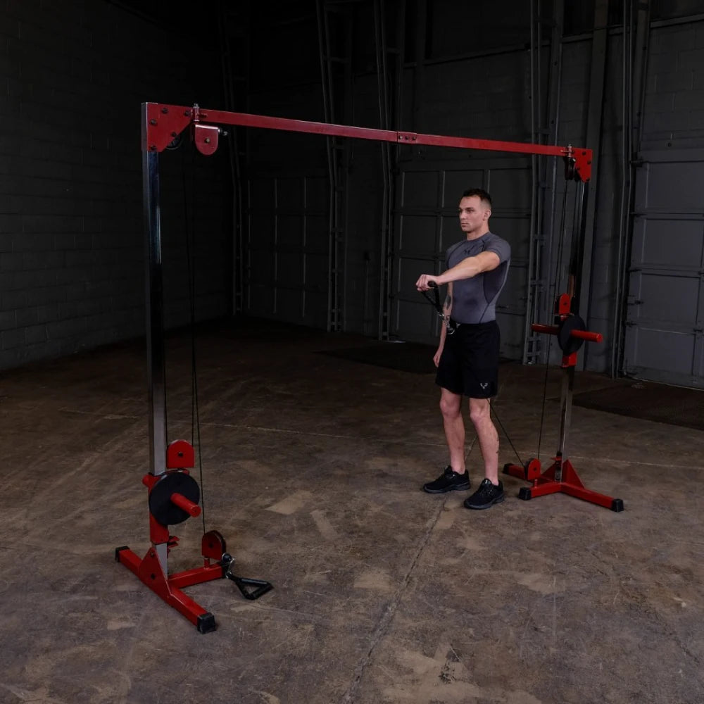 A man in a fitted gray shirt and black shorts stands next to a cable machine in a gym, preparing to perform an exercise. The machine features a red and black design, with cables hanging down. The gym has a concrete floor and a simple backdrop.
