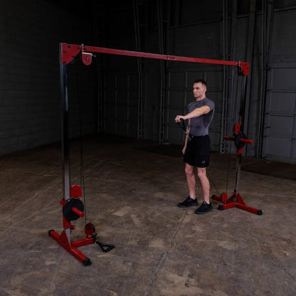 A man in a fitted gray shirt and black shorts stands next to a cable machine in a gym, preparing to perform an exercise. The machine features a red and black design, with cables hanging down. The gym has a concrete floor and a simple backdrop.