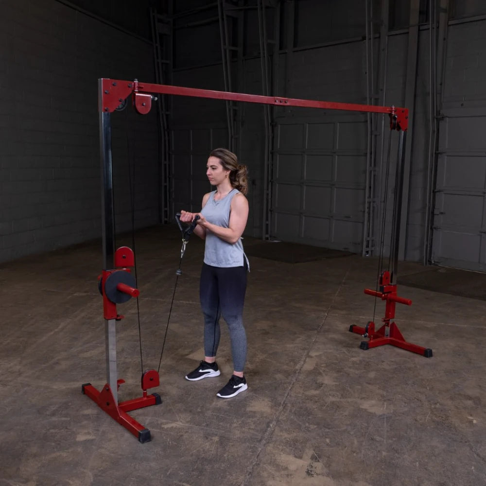 A woman stands in a gym setting, holding the handles of a cable machine, preparing to perform an exercise. She is wearing a light gray tank top and dark leggings, with her hair styled in loose waves. The gym features a red and silver cable machine against a plain wall.