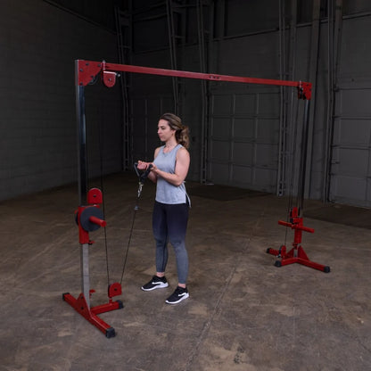 A woman stands in a gym setting, holding the handles of a cable machine, preparing to perform an exercise. She is wearing a light gray tank top and dark leggings, with her hair styled in loose waves. The gym features a red and silver cable machine against a plain wall.