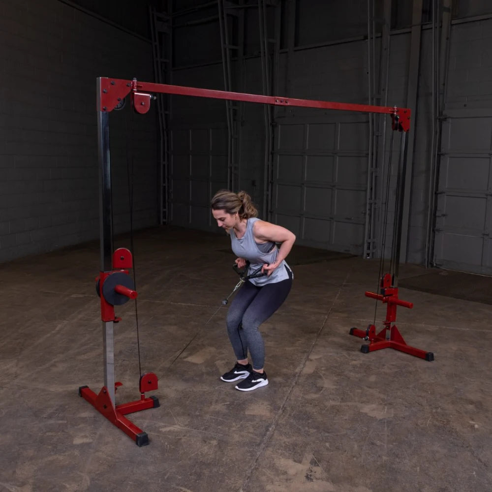A woman performing a seated cable row exercise using a red and silver cable machine in a spacious gym setting with a concrete floor. She is wearing a gray tank top and black leggings, focusing on her form while pulling the cable towards her.