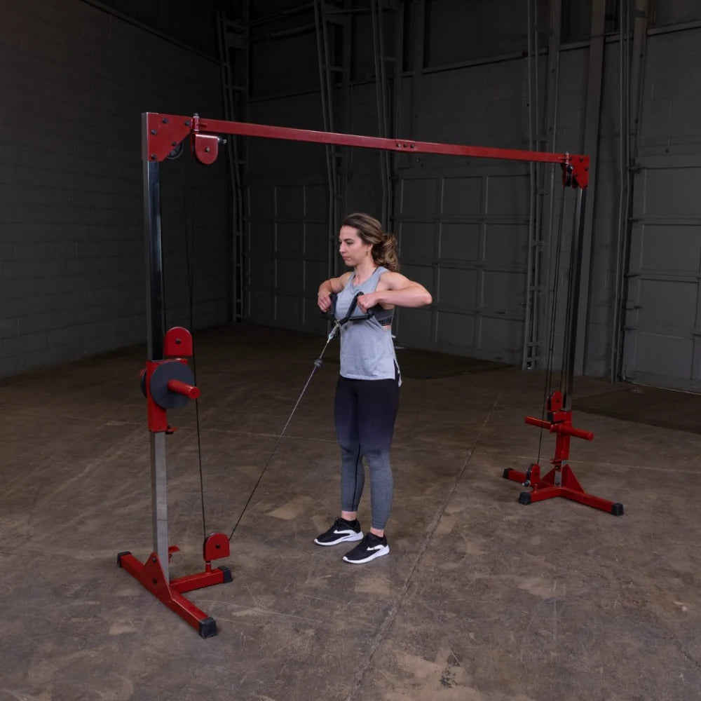 A woman performs a cable exercise using a red and silver cable machine in a gym setting, wearing a gray tank top and black leggings.