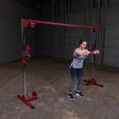 A woman performs an exercise using a cable machine in a gym setting, wearing a light gray tank top and black leggings, with her hair styled in loose waves. The background features a spacious gym area with gray walls and a concrete floor.