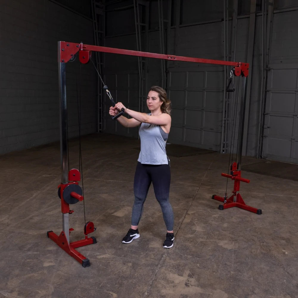 A woman performs a cable exercise using a red cable machine in a gym setting, wearing a light gray tank top and black leggings.